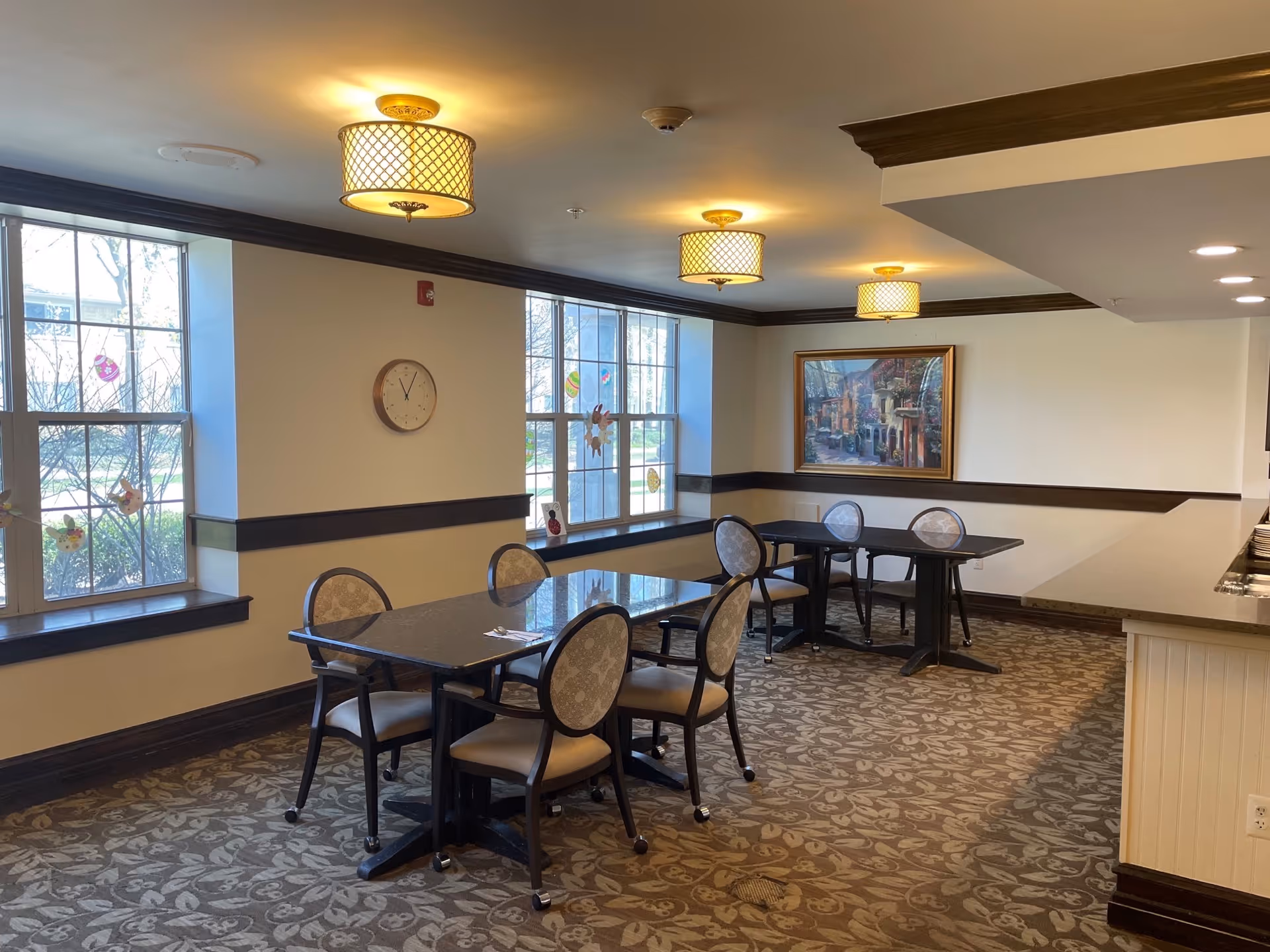 A dining area in Chester Street Residence with two tables, each surrounded by four cushioned chairs. The room has large windows with decorative stickers, a wall clock, and a framed painting. The ceiling features three round light fixtures, and the floor is carpeted with a floral pattern.
