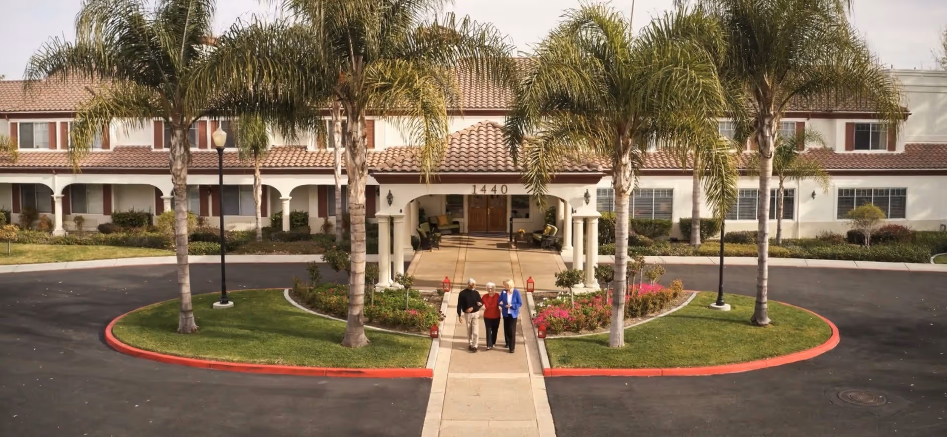 Front exterior view of Aegis Living Shadowridge facility with a driveway, palm trees, and three elderly people walking on the sidewalk towards the entrance under a covered porch.