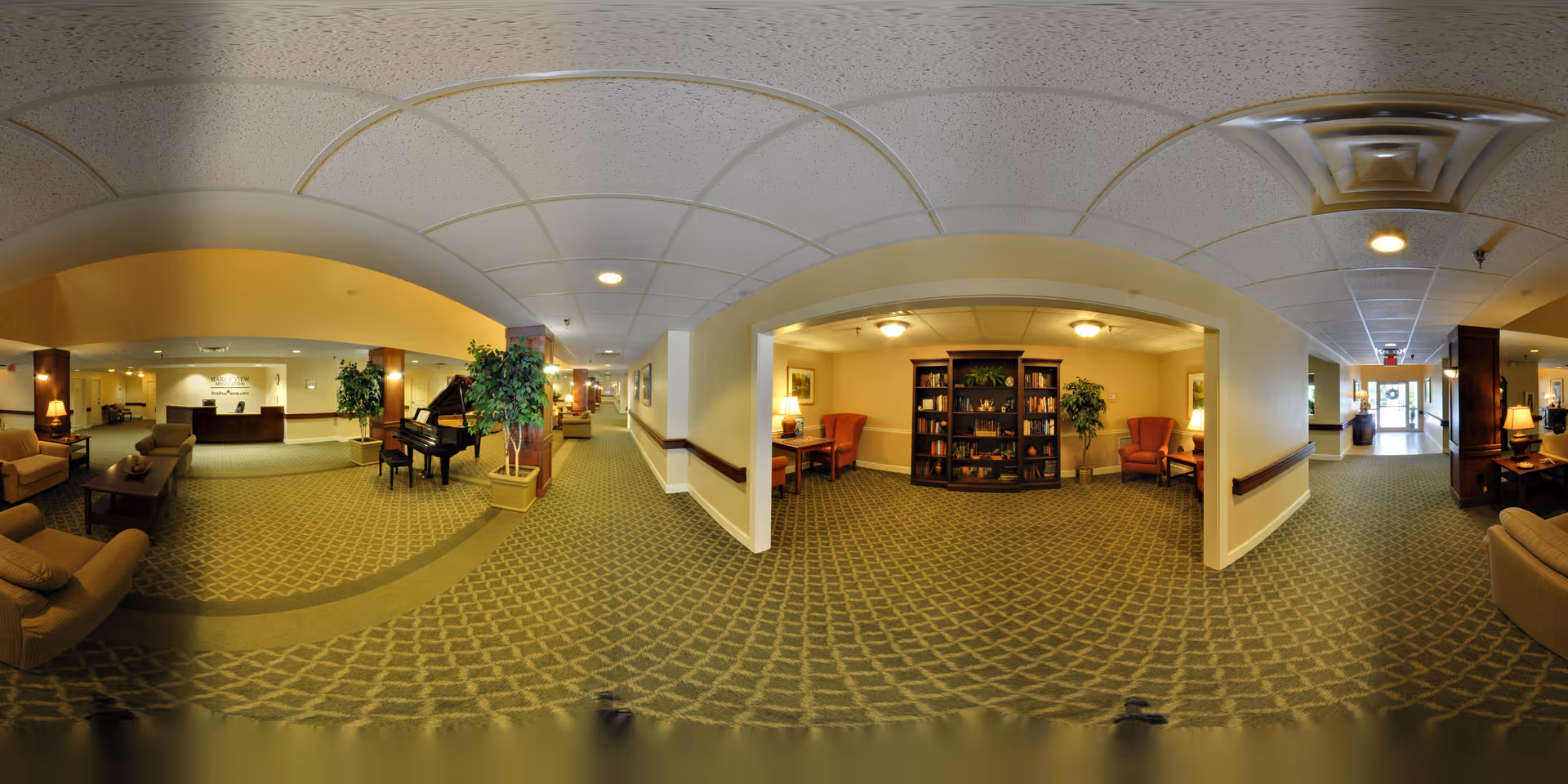 A spacious senior living facility interior with a carpeted floor and a patterned design. The area includes comfortable seating with armchairs and sofas, a grand piano near a pillar with a potted plant, a reception desk in the background, and a cozy reading nook with a bookshelf and two armchairs. The ceiling has recessed lighting and ventilation panels, and the hallway extends toward an entrance with glass doors.