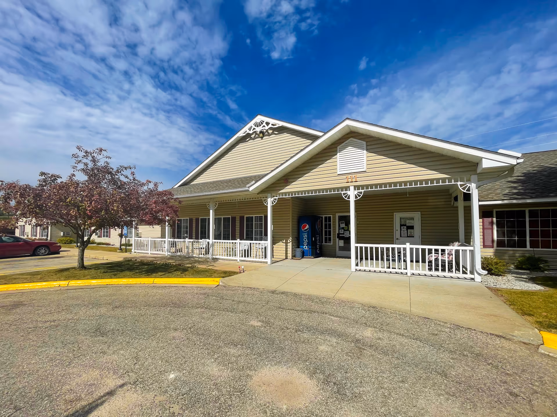 Exterior view of a single-story beige building with white trim and a covered porch area. There is a Pepsi vending machine near the entrance, a tree with red leaves on the left, and a red car parked nearby. The sky is partly cloudy with blue patches.