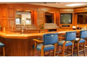 Interior bar area with a wooden counter, blue upholstered bar stools, glassware, and built-in cabinetry with a TV.