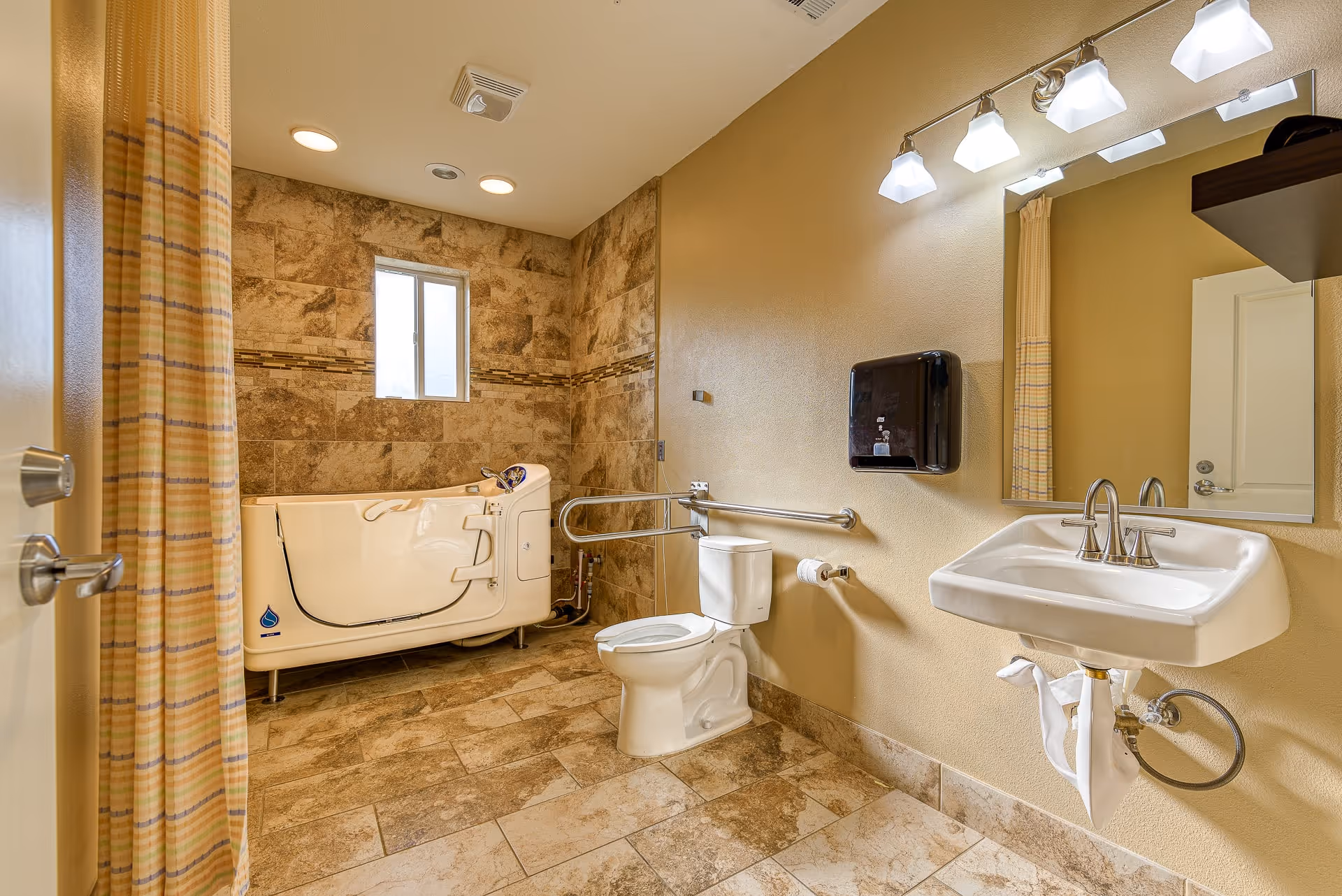 Accessible bathroom with a walk-in bathtub, toilet, wall-mounted sink, grab bars, and tiled walls and floor.