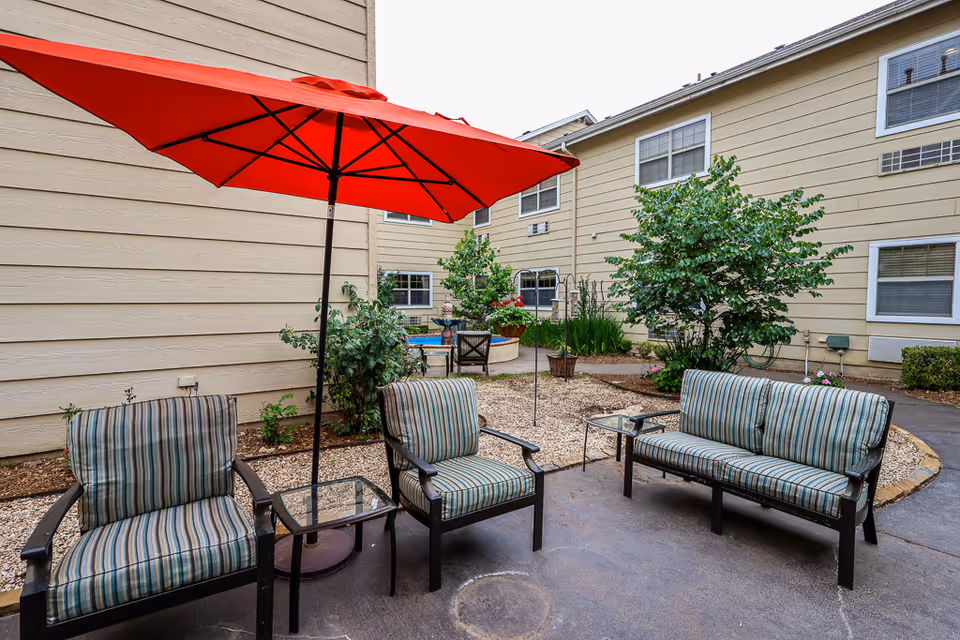 Outdoor seating area with striped cushioned chairs and a loveseat under a large red umbrella, surrounded by beige building walls and greenery.
