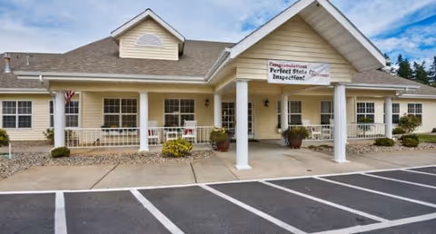 Exterior front view of a single-story senior living facility building with a covered entrance supported by white columns. The building has beige siding, multiple windows, and a banner hanging above the entrance that reads 'Congratulations Perfect State Inspection!'. There are potted plants near the entrance and a parking lot with marked spaces in front.