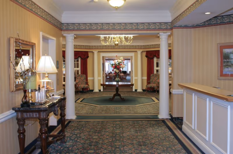 Interior lobby with columns, a central flower-topped table, reception desk, and patterned carpet and wallpaper.