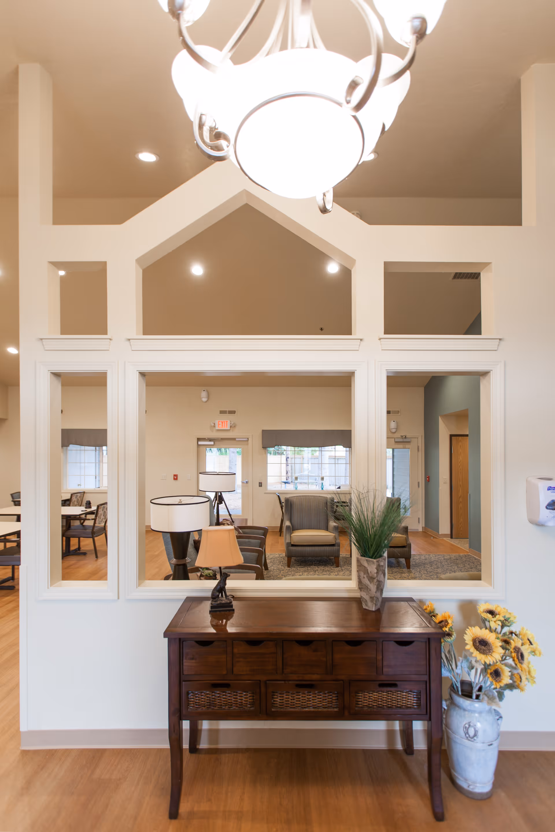 Interior view of a senior living facility showing a wooden console table with drawers and baskets, a small lamp, and a vase with green plants on top. To the right of the table is a large ceramic vase with sunflowers. Behind the table is a large window opening into a common area with chairs, tables, and lamps. The walls are light-colored and the floor is wooden.