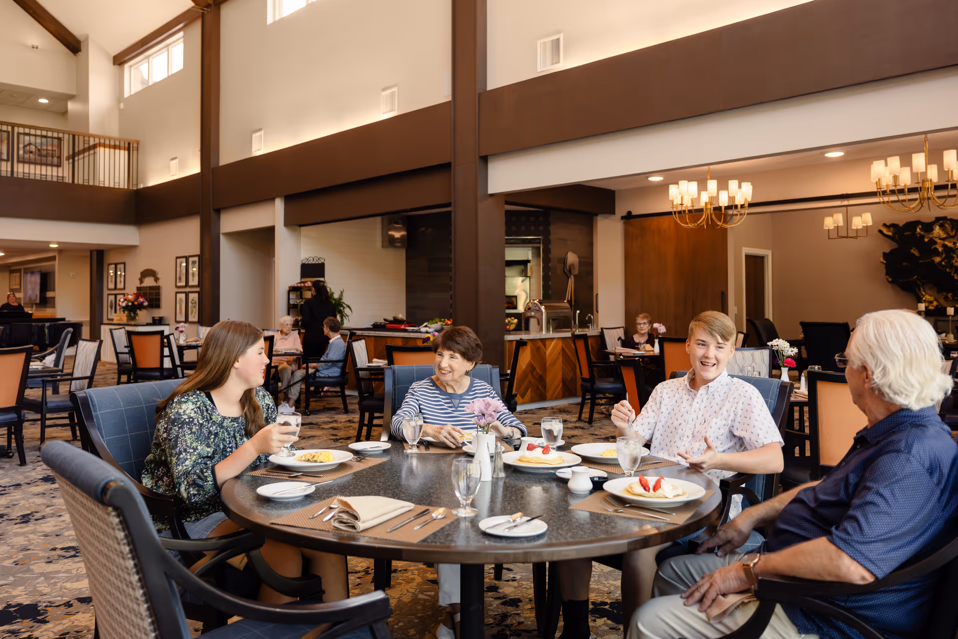 A group of four people, including two elderly adults and two younger individuals, sitting around a round dining table in a spacious, well-lit dining room. They are enjoying a meal together, with plates of food and glasses of water on the table. The room features high ceilings, large windows, chandeliers, and a warm, inviting atmosphere with other diners visible in the background.