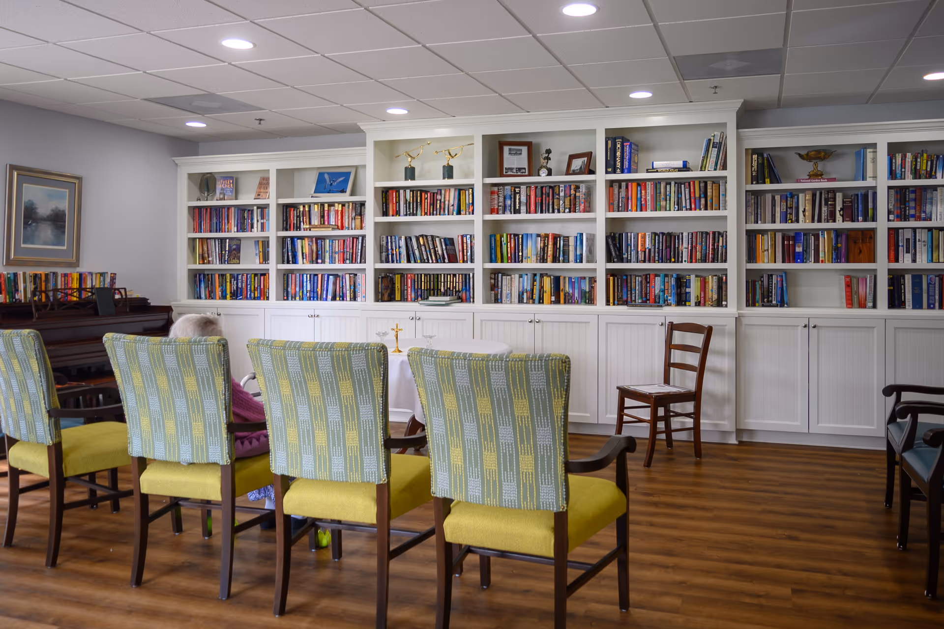 A cozy library or common room with a row of green and yellow patterned chairs facing a white bookshelf filled with books. There is a piano on the left side and a wooden chair on the right. The room has wood flooring and a ceiling with recessed lighting.