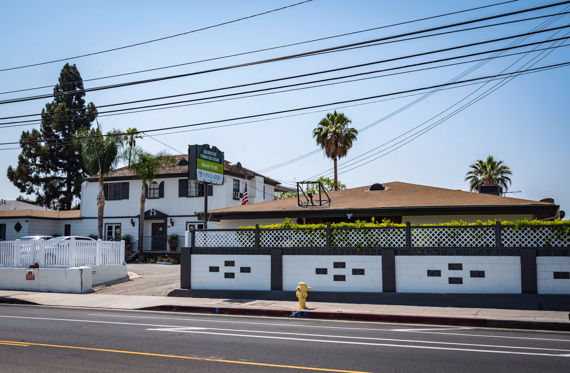 Street-facing view of a two-story post-acute care building behind a white decorative wall and fence with palm trees and a fire hydrant in front.