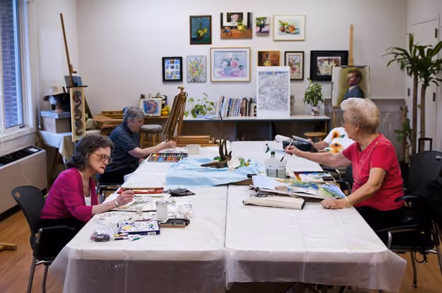 Three elderly women seated around a large table in an art room, painting and drawing. The table is covered with a white plastic sheet and art supplies including paint, brushes, and paper are spread out. The room has wooden floors, several framed artworks on the wall, and easels with paintings in the background.