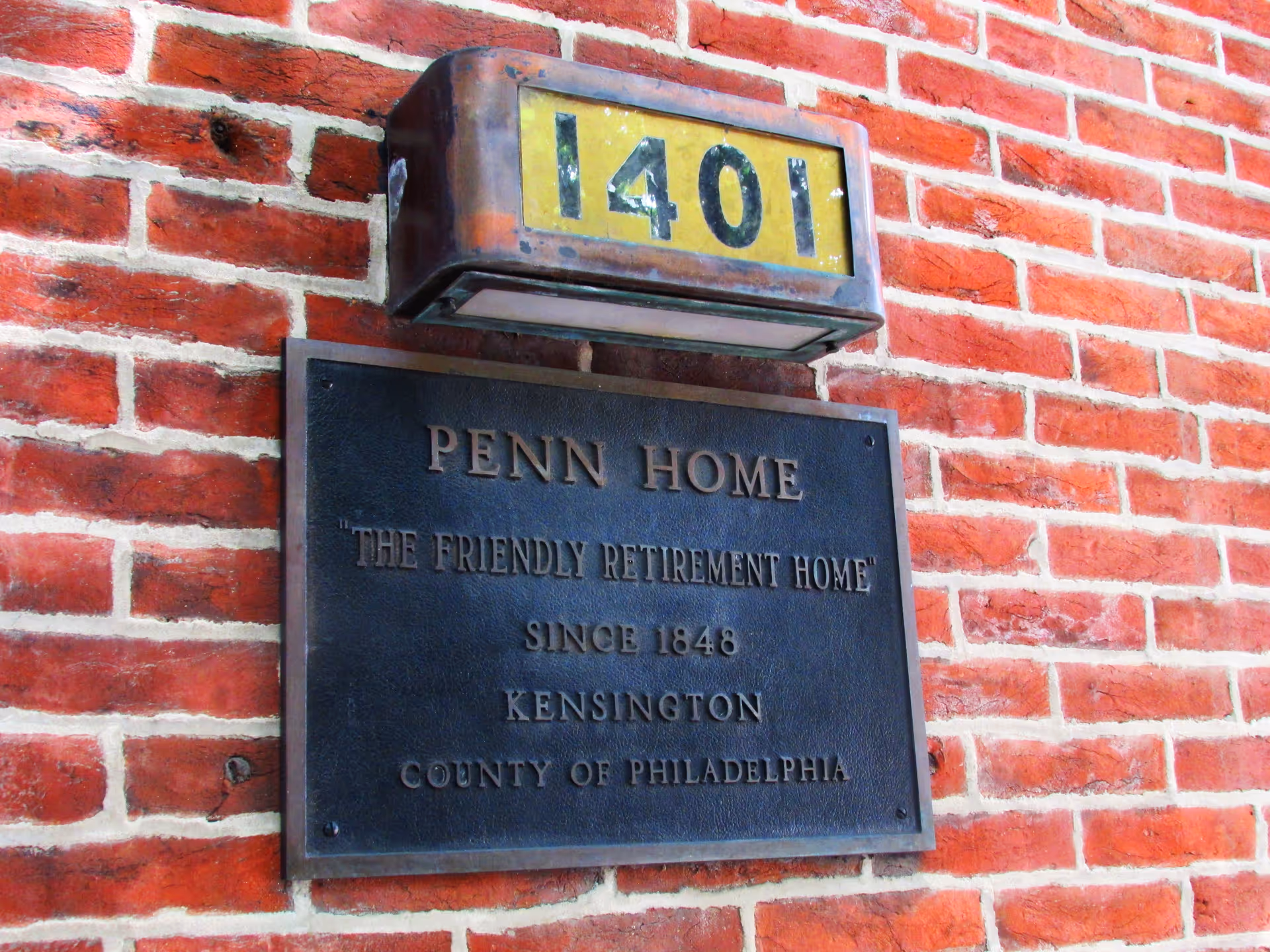A close-up view of a brick wall with a metal plaque and a vintage address light above it. The plaque reads: 'PENN HOME "THE FRIENDLY RETIREMENT HOME" SINCE 1848 KENSINGTON COUNTY OF PHILADELPHIA'. The address light above displays the number 1401.