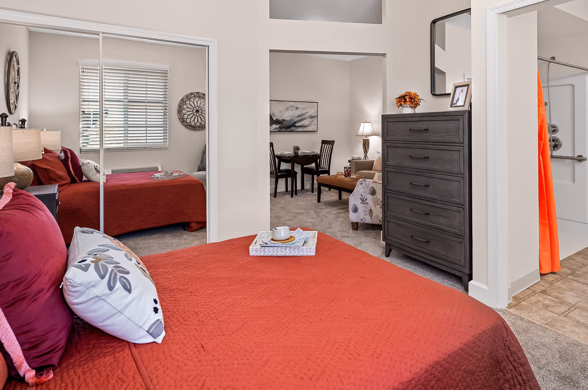 Interior view of a senior living facility room featuring a bed with red bedding and decorative pillows, a dark wooden dresser, and a small living area with a table and chairs in the background. A bathroom with a shower and orange curtain is visible to the right.