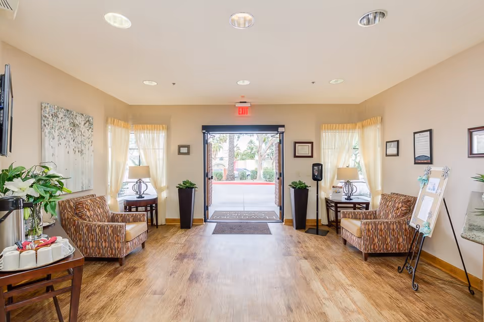 Bright and welcoming lobby area of a senior living facility with two patterned armchairs on either side, small side tables with lamps, potted plants, framed artwork and certificates on the walls, a coffee station with cups and flowers on the left, and an open glass door leading outside with an exit sign above it.