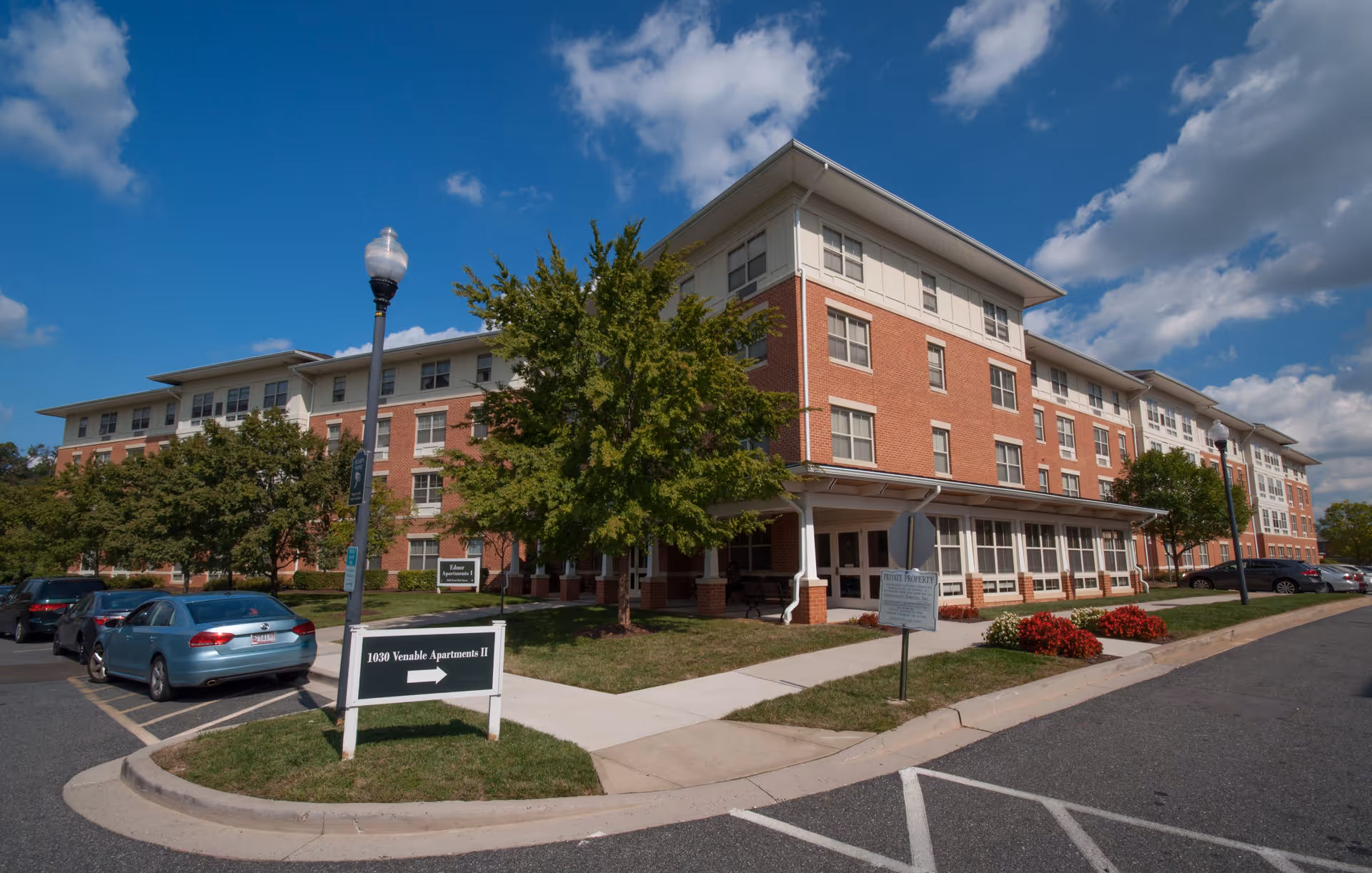 Exterior view of a multi-story brick and white-paneled apartment building under a blue sky with scattered clouds. There are trees and parked cars along the sidewalk, and signs indicating directions to 1030 Venable Apartments II and Ednor Apartments.