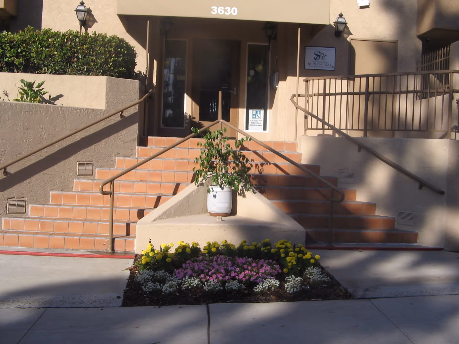 Entrance to a building with beige walls and terracotta tiled stairs. There are handrails on both sides of the stairs and a ramp in the middle with a potted plant. In front of the stairs is a small garden bed with yellow, pink, and white flowers. The building number 3630 is displayed above the entrance.