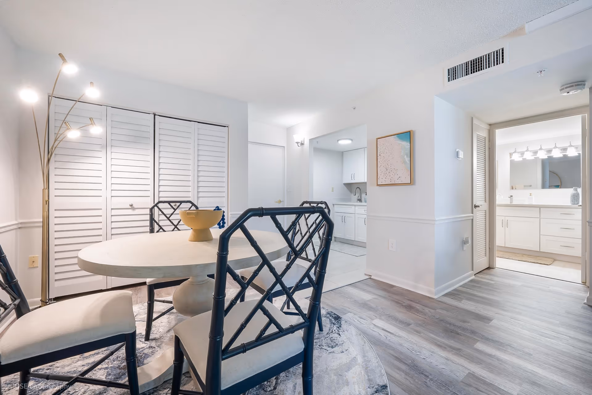 Light-filled dining area with a round table and four chairs adjacent to a kitchen and bathroom in a senior living unit.