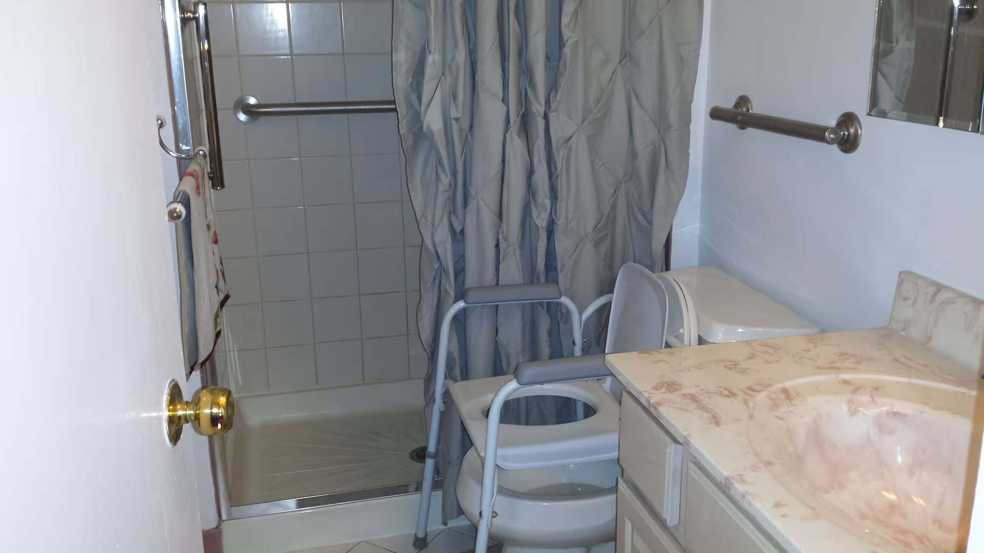A small bathroom with a shower area that has a grab bar and a shower curtain. There is a raised toilet seat with armrests in front of the toilet. A towel hangs on a rack attached to the door, and a sink with a marble-patterned countertop is visible on the right side.