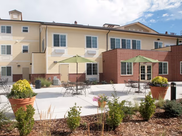 Outdoor patio area of a senior living facility with several metal tables and chairs, each shaded by green umbrellas. The building exterior is a combination of yellow siding and red brick, with multiple windows and a clear sky above. There are large potted plants and landscaped greenery in the foreground.