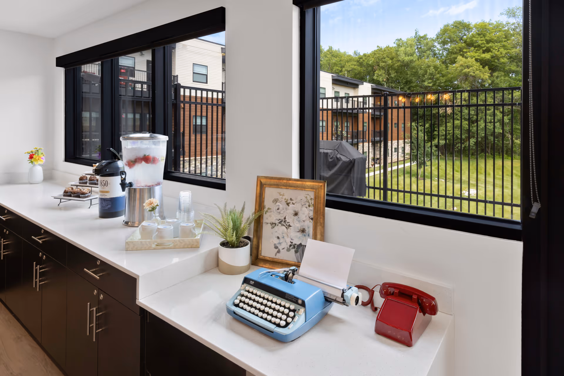 A bright interior countertop area with large windows overlooking a fenced grassy outdoor space and neighboring buildings. On the countertop are a blue vintage typewriter with a sheet of paper, a red rotary telephone, a framed floral picture, a small potted plant, a water dispenser with infused water, cups, a coffee dispenser, and a tray with pastries. The cabinetry below is dark with silver handles.