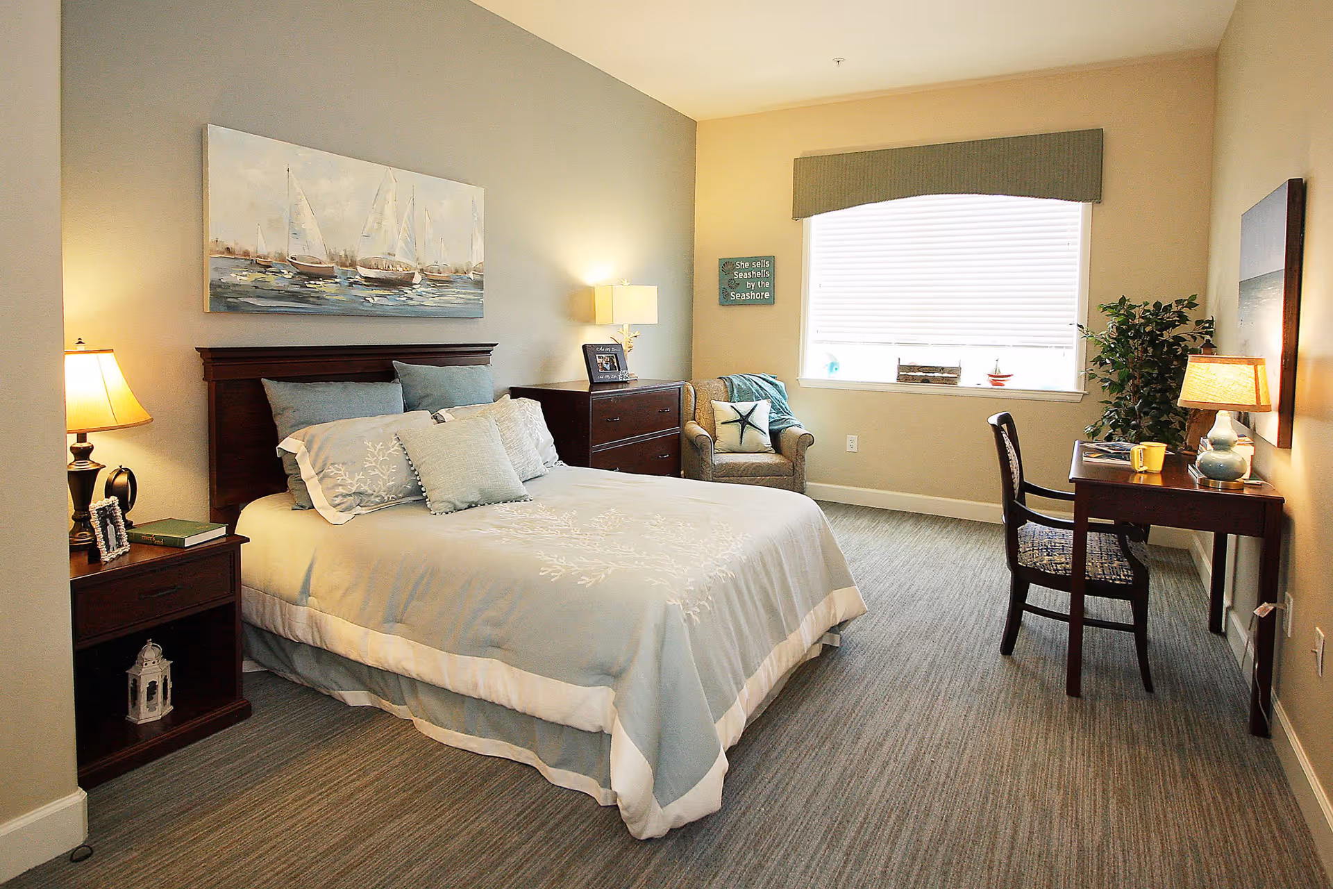 A cozy bedroom with a neatly made bed featuring light blue and white bedding. The room has a wooden headboard, two bedside tables with lamps, a dresser, an armchair with a decorative pillow, a desk with a chair and lamp, and a large window with blinds and a valance. The walls are painted in neutral tones and decorated with paintings and a small sign.