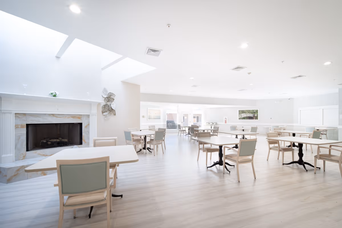 A bright and spacious dining room with multiple tables and chairs arranged neatly. The room features light-colored flooring, a white ceiling with recessed lighting, and a decorative fireplace on the left side. Large windows and glass doors at the far end allow natural light to fill the space.