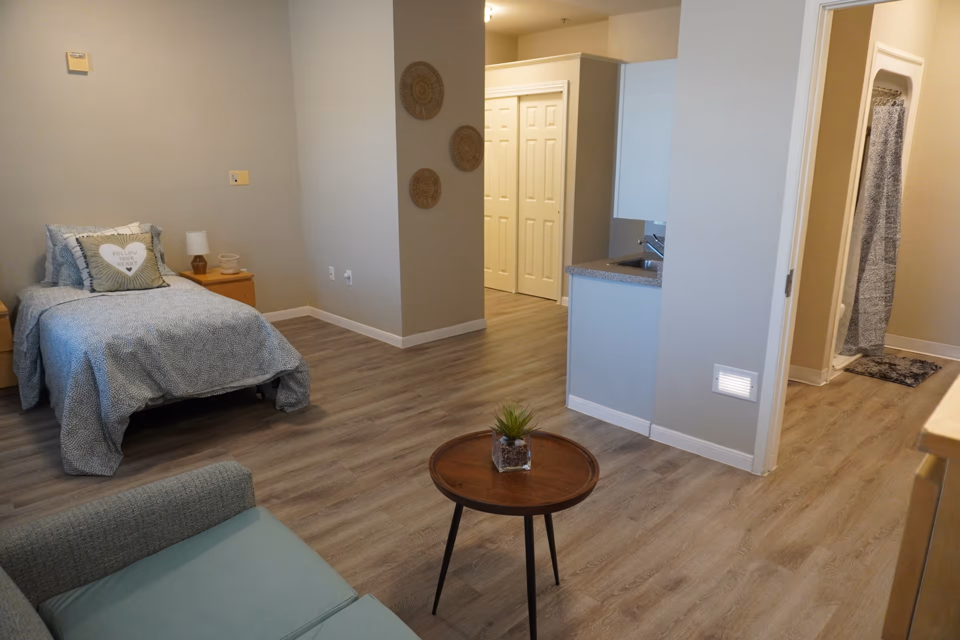 A senior living facility room with a single bed dressed in blue patterned bedding and a decorative pillow, a small wooden nightstand with a lamp, a light blue armchair, a round wooden coffee table with a small plant, and a kitchenette area with a sink. The room has wood-style flooring and light-colored walls, with a bathroom visible in the background featuring a shower with a curtain.