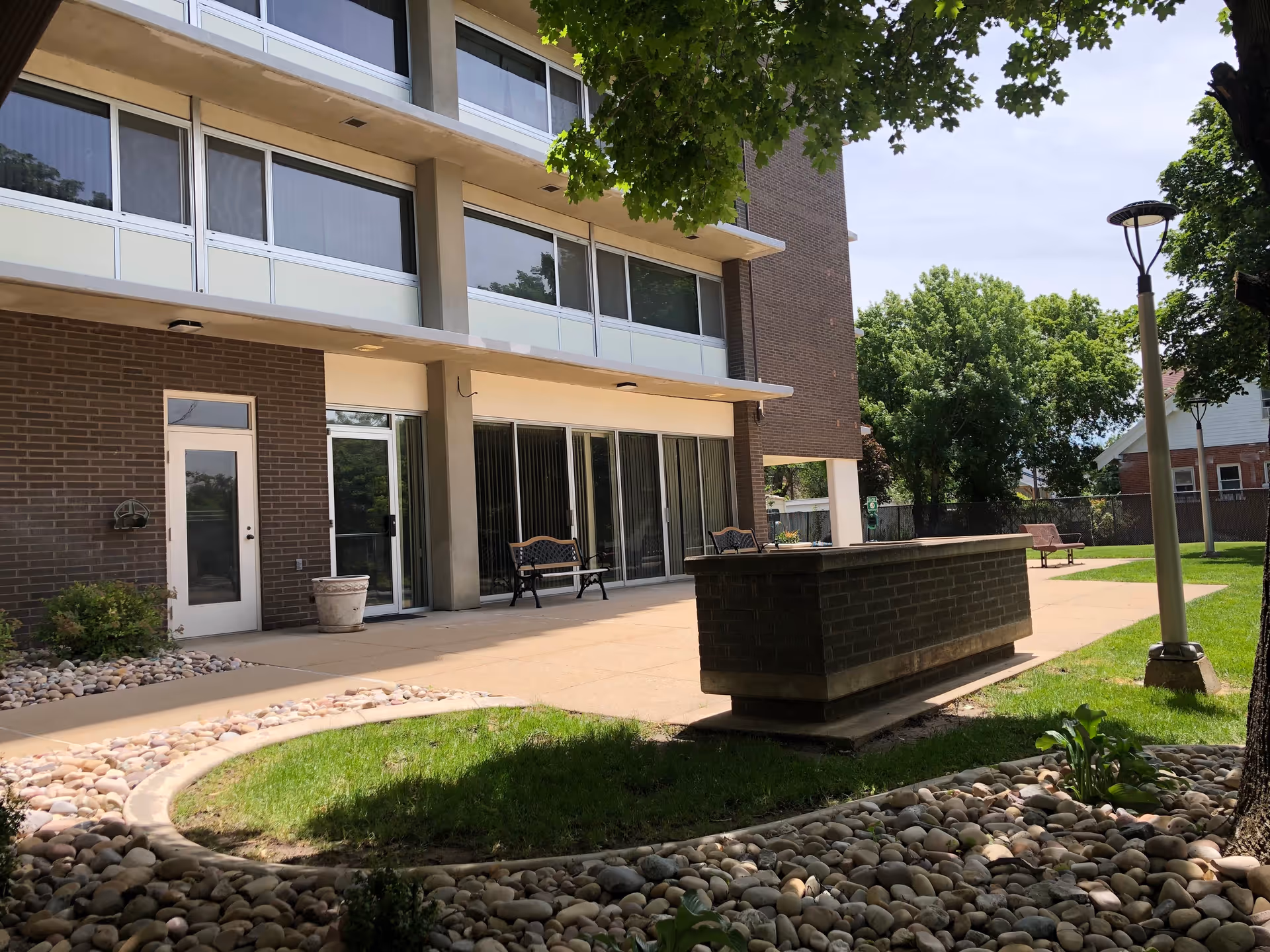 Exterior courtyard and patio of a multi-story brick senior living building with benches, planters, and trees.