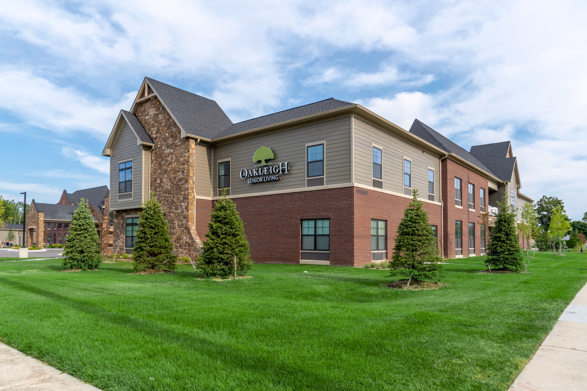 Exterior view of Oakleigh of Macomb Senior Living building with a combination of brick and stone facade, surrounded by green grass and small trees under a partly cloudy sky.