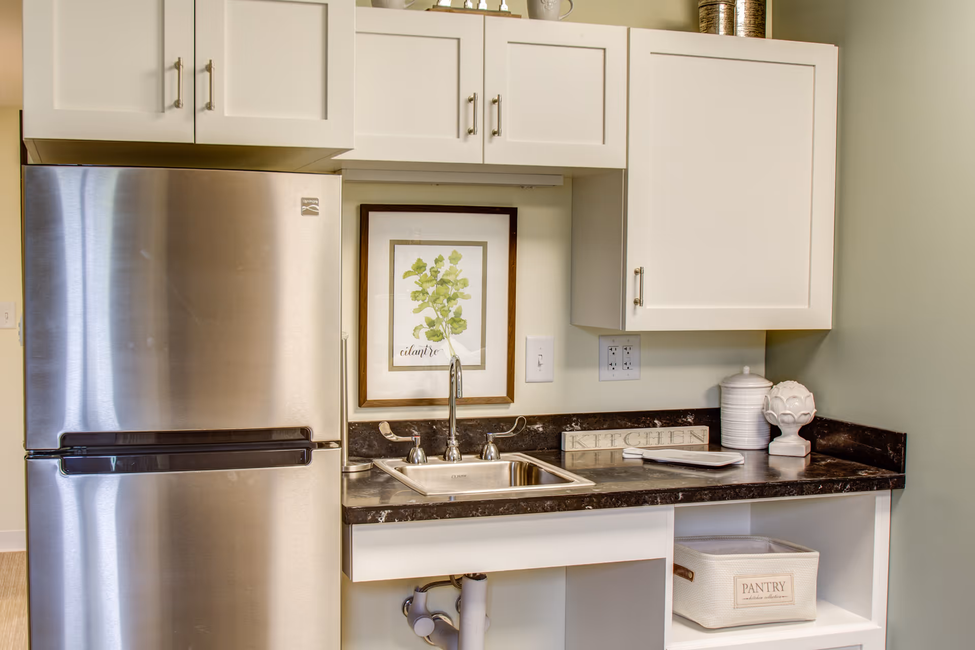 A small kitchen area featuring a stainless steel refrigerator, white cabinets, a dark countertop with a built-in sink, and decorative items including a framed cilantro plant illustration, a white canister, and a sign that says 'KITCHEN'. Below the counter is an open shelf with a white basket labeled 'PANTRY'.