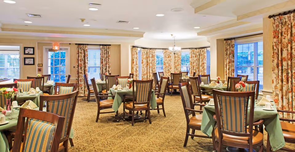 A dining room with multiple round tables covered with green tablecloths, each set with napkins, cups, and small flower arrangements. The room has wooden chairs with striped upholstery, floral curtains on the windows, and a carpeted floor with a patterned design. Ceiling lights and a chandelier provide illumination.
