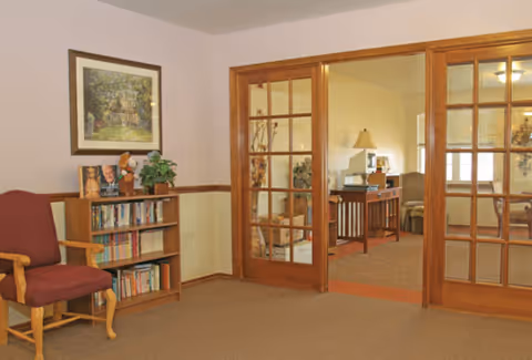 A cozy interior room with beige walls and carpeted floor. There is a wooden bookshelf filled with books and a few decorative items, including a small plant and a teddy bear. A framed picture hangs above the bookshelf. To the right, wooden framed glass double doors open into another room with a table, lamp, and seating area visible.