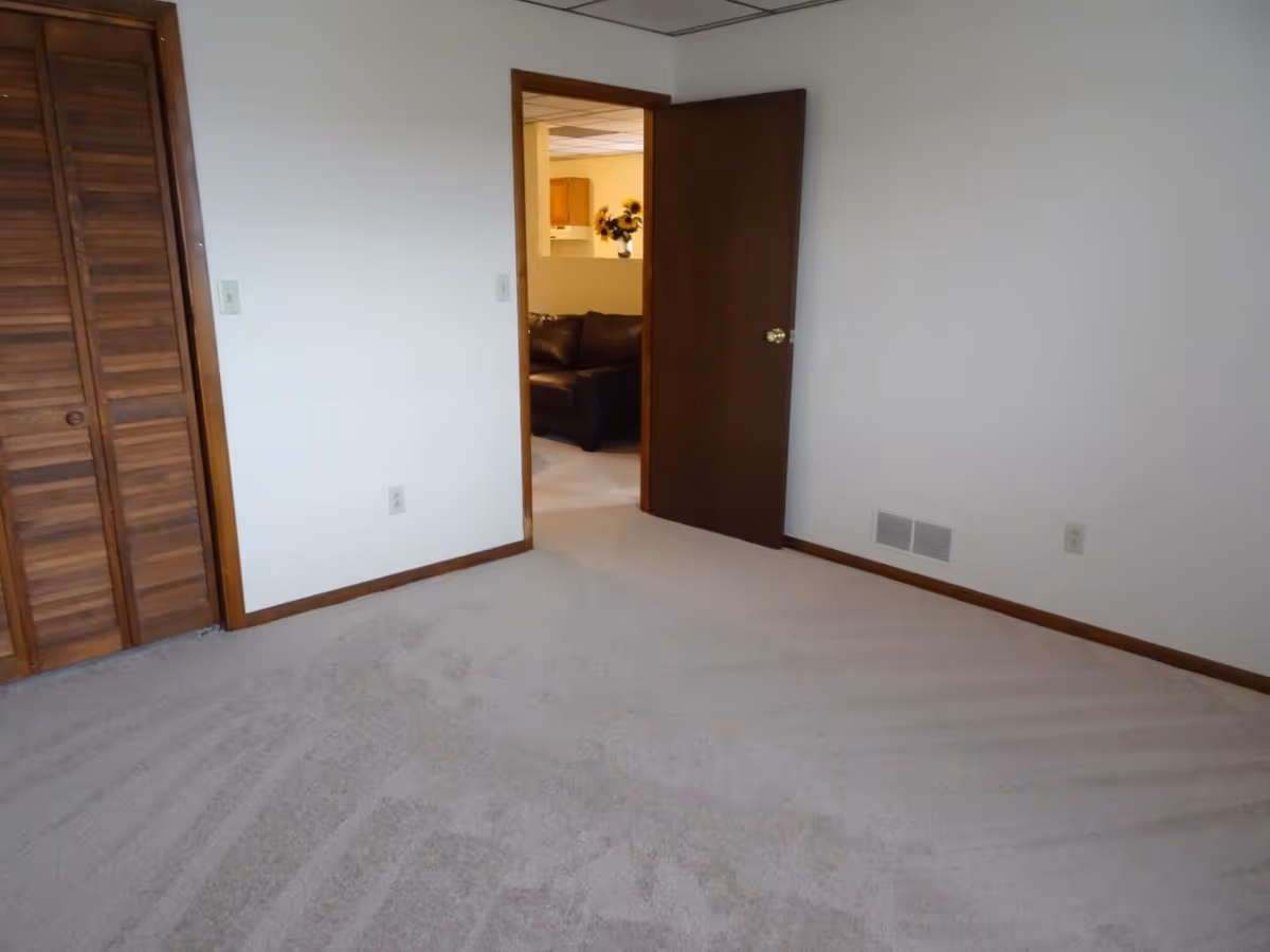 Empty carpeted bedroom with louvered wooden closet doors and an open interior door showing a couch in the next room.