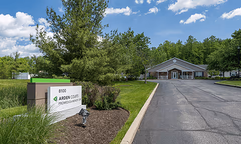 Exterior view of Arden Courts - ProMedica Memory Care Community in Chagrin Falls, showing a driveway leading to the building entrance surrounded by green trees and grass under a partly cloudy sky. A sign with the facility name and address is visible near the road.