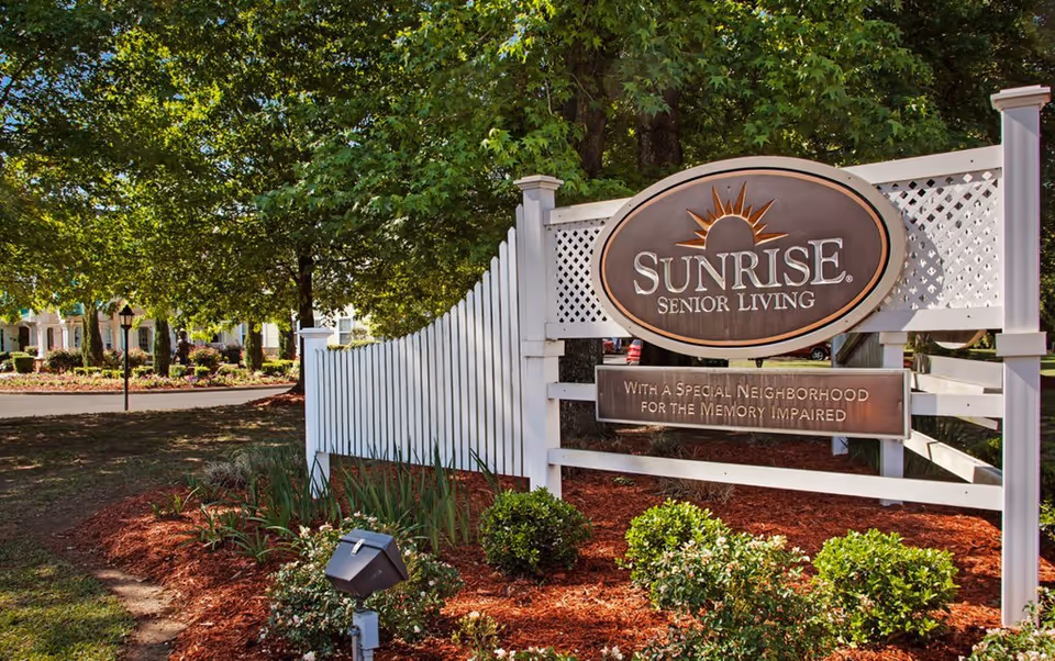 Outdoor view of a Sunrise Senior Living sign mounted on a white wooden fence with greenery and trees in the background. The sign reads 'Sunrise Senior Living' and 'With a Special Neighborhood for the Memory Impaired.'