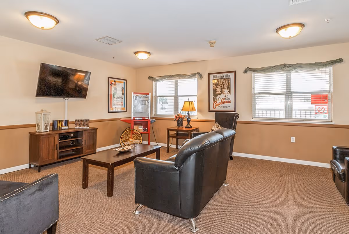 A cozy living room with beige walls and carpeted floor featuring a black leather sofa, a matching armchair, a wooden coffee table, and a wooden TV stand with a flat-screen TV mounted above it. There are two windows with blinds and green valances, a side table with a lamp and flowers, a red popcorn machine, and framed vintage movie posters on the walls.