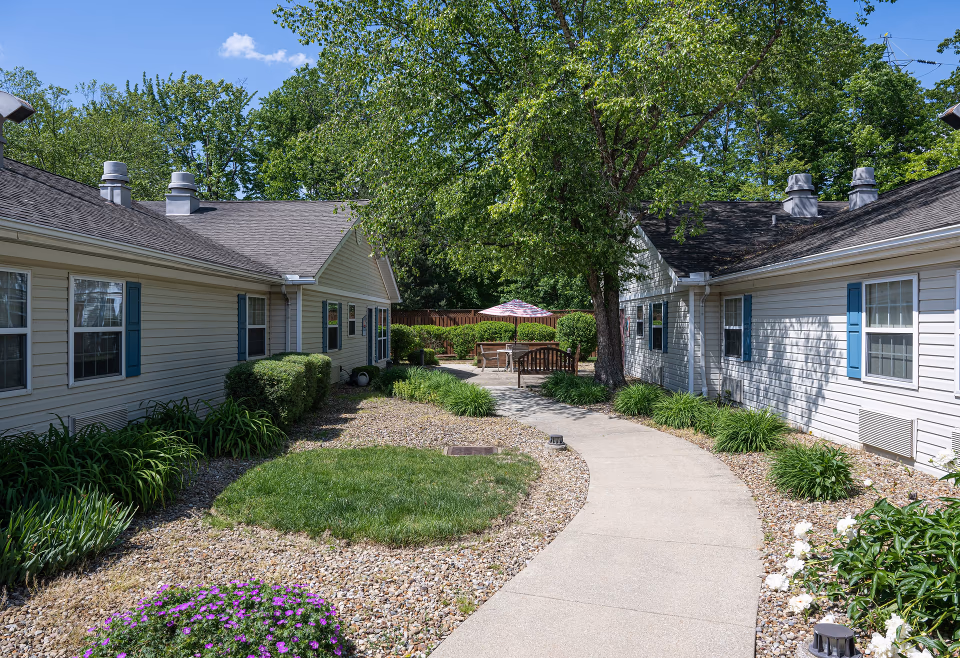 Outdoor courtyard area between two single-story buildings with white siding and blue shutters. A curved concrete pathway leads to a seating area with a table, chairs, and an umbrella. The courtyard is landscaped with green bushes, grass, flowers, and trees under a clear blue sky.