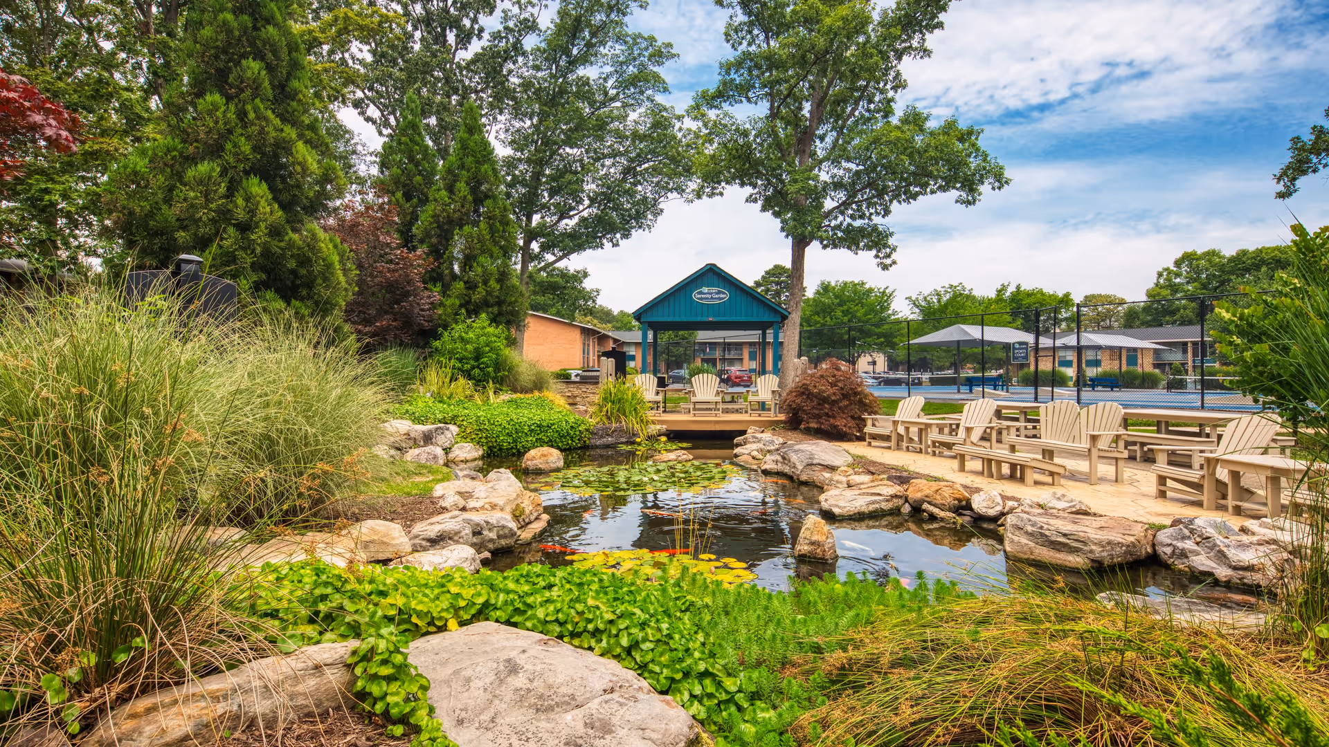 A serene garden area featuring a pond with lily pads, surrounded by lush greenery and rocks. In the background, there is a blue building with a porch and several wooden chairs, along with a tennis court visible behind a fence.