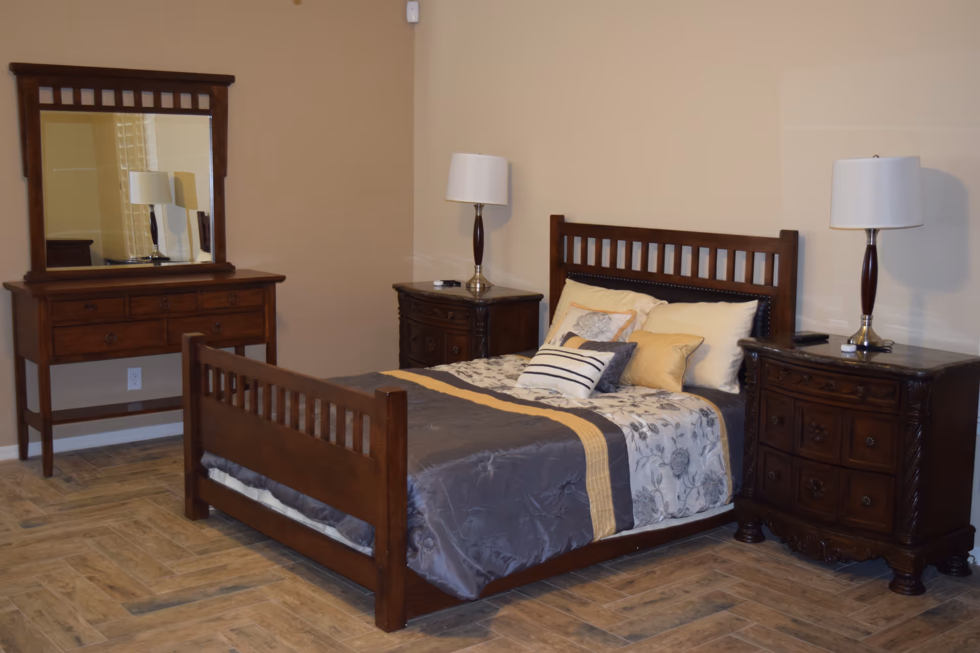 A bedroom with a wooden bed frame, a bed made with gray, yellow, and floral patterned bedding, two wooden nightstands each with a lamp, and a wooden dresser with a large mirror. The floor has a wood-like tile pattern and the walls are painted beige.