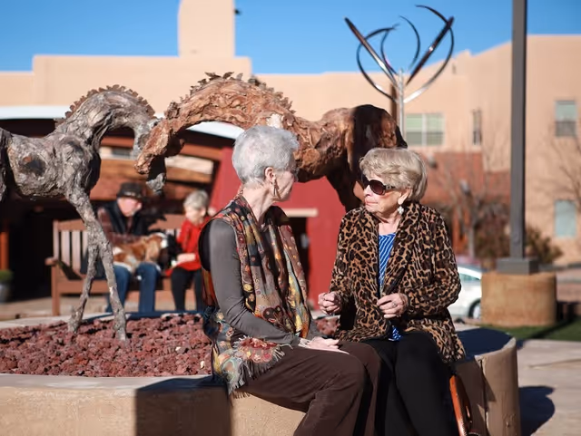 Two elderly women sitting and talking outdoors near a sculpture of two horses. One woman has short gray hair and wears glasses and a patterned scarf, while the other woman wears sunglasses and a leopard print jacket. In the background, two other people are sitting on a bench with a dog, and a building with adobe-style architecture is visible.
