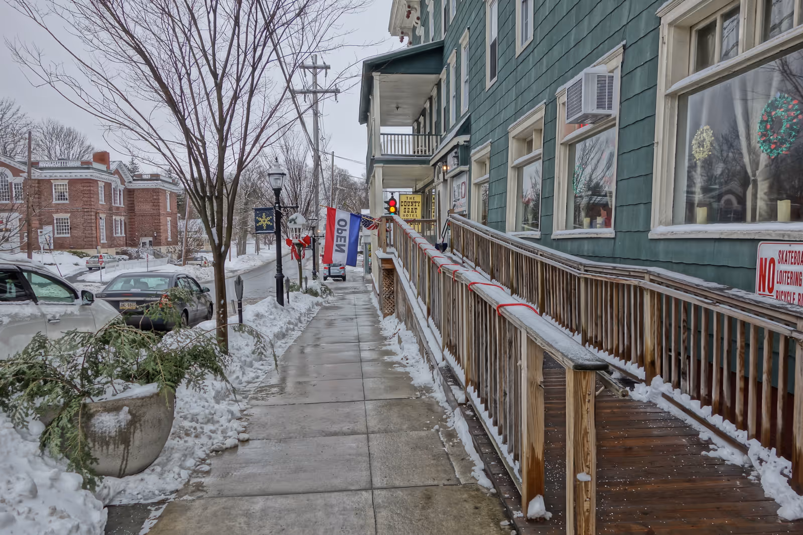 Sidewalk covered with light snow next to a green building with a wooden ramp and several windows decorated with holiday wreaths. There are parked cars along the street, leafless trees, and a flag with the word 'OPEN' hanging near the building entrance. A sign for 'The County Seat Hotel' is visible in the background.