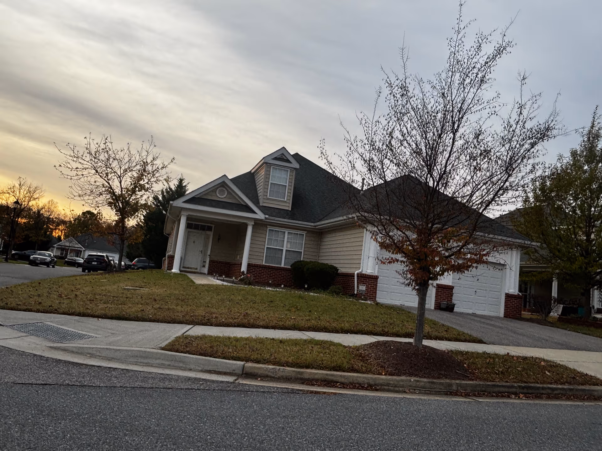 A single-story residential house with beige siding and a brick foundation, featuring a front porch with white columns, a dormer window on the roof, and a two-car garage. There is a small tree with sparse leaves in the front yard and a sidewalk along the street. The sky is cloudy with the sun setting in the background.