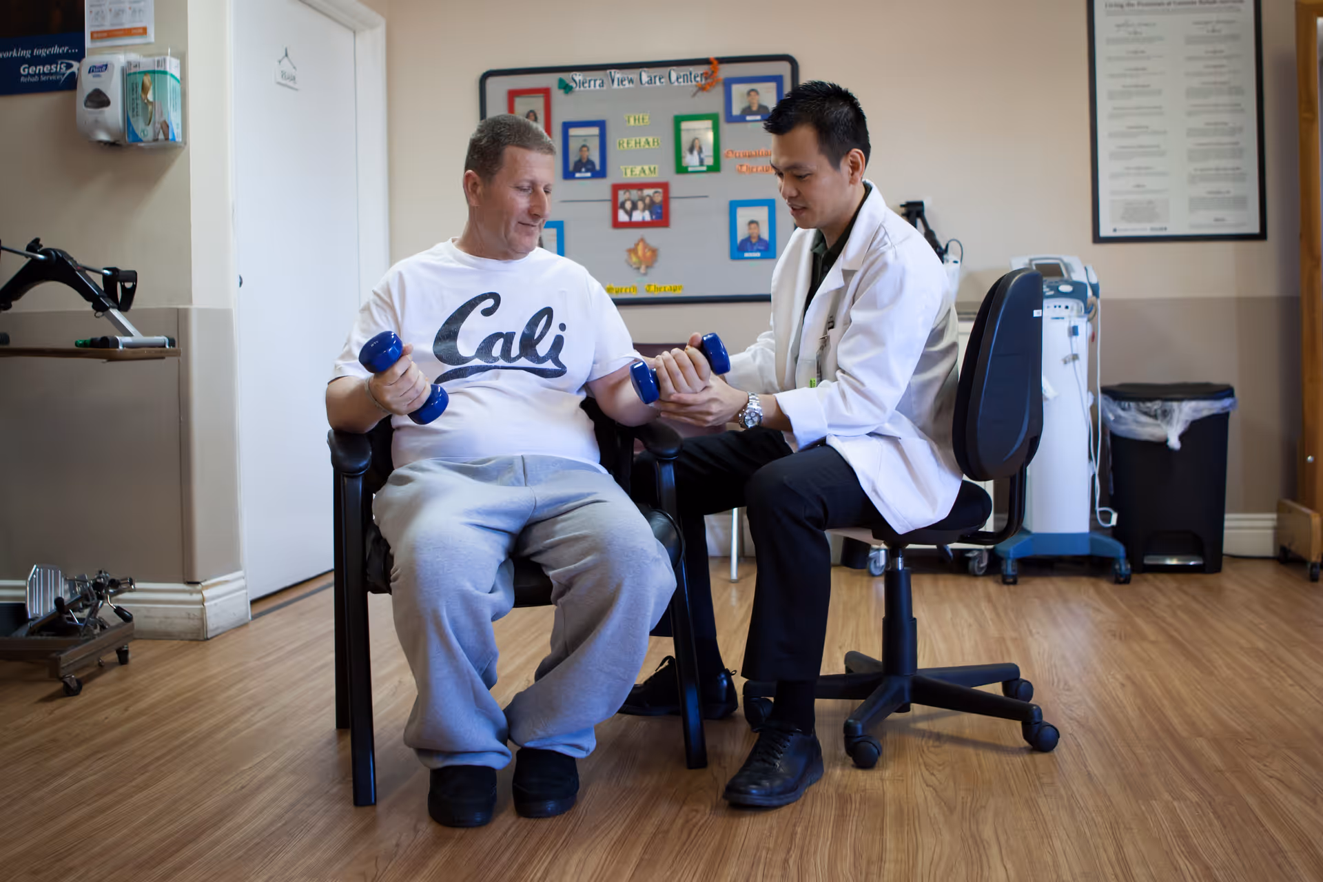 A man in casual clothes holding blue dumbbells is seated in a chair while a healthcare professional in a white coat assists him with arm exercises in a room with wooden flooring and medical equipment.
