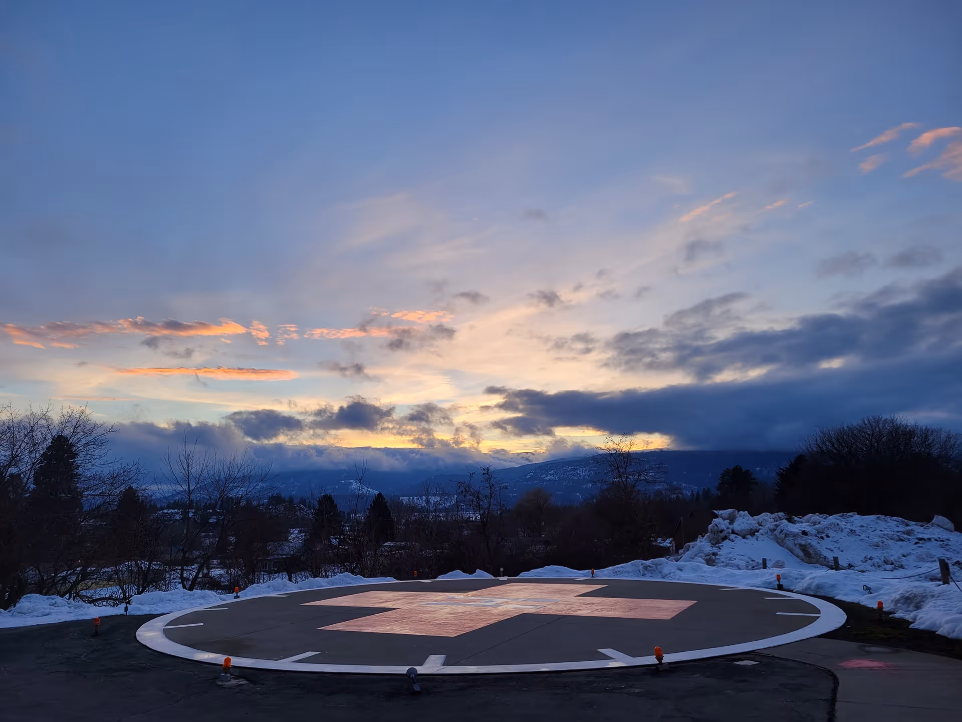 Circular helipad at dusk with snow piles, trees and distant mountains beneath a colorful sunset sky.