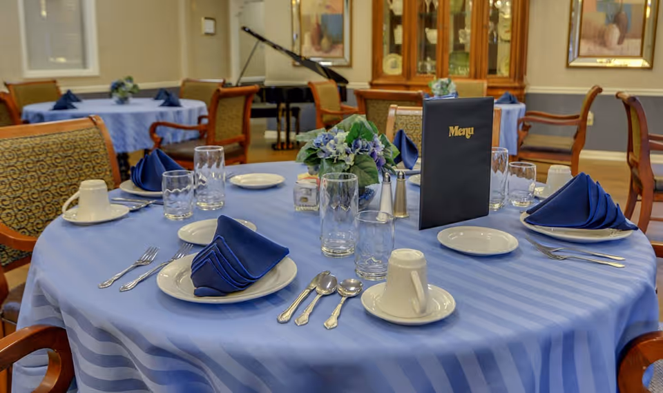 A dining table set with a blue striped tablecloth, blue folded napkins on white plates, silverware, glasses, coffee cups, a floral centerpiece, salt and pepper shakers, and a menu standing upright. In the background, more similarly set tables and a grand piano are visible in a well-lit room.