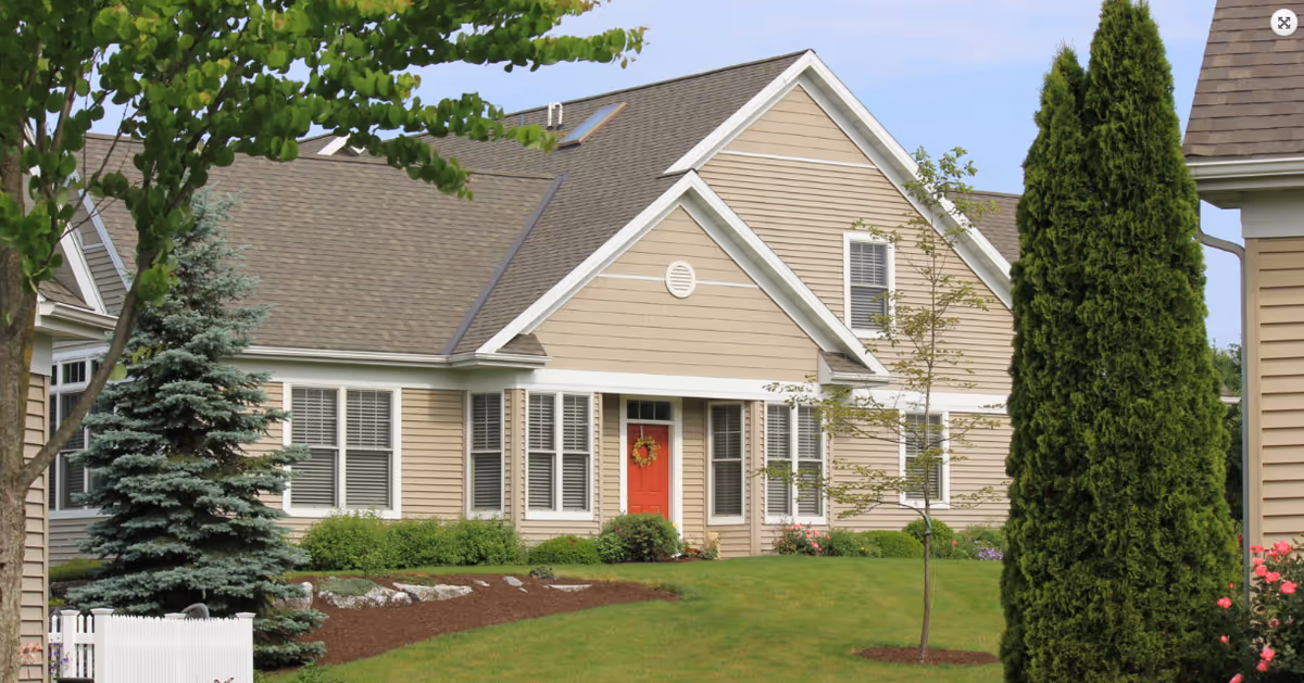 Front exterior of a beige single-story residential building with a red door, manicured lawn, and surrounding trees and shrubs.