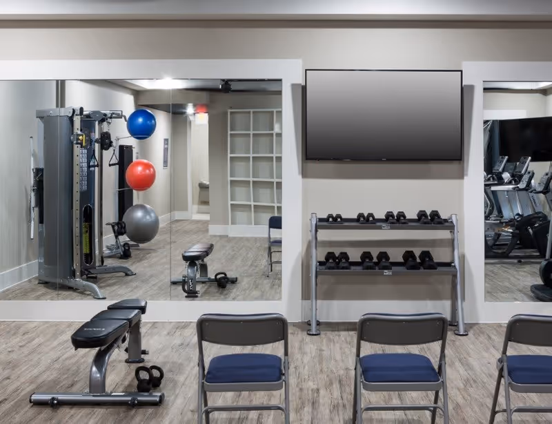 Interior view of a fitness room with exercise equipment including a weight bench, dumbbells on a rack, kettlebells, and exercise balls. Large mirrors cover the walls, reflecting the equipment and space. A flat-screen TV is mounted on the wall above the dumbbell rack. Three chairs with blue cushions are positioned facing the TV and equipment.