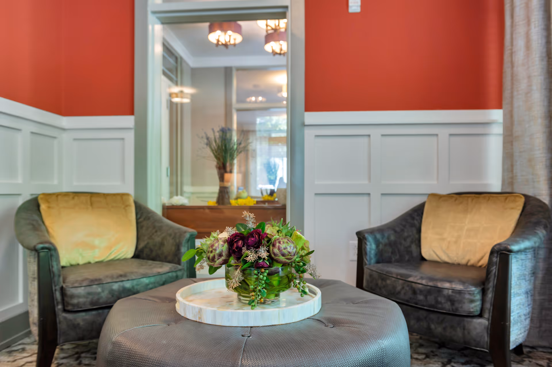A cozy sitting area with two dark armchairs each with a yellow cushion, a round ottoman with a tray holding a green vase filled with flowers, red and white paneled walls, and a mirror reflecting a hallway with a table and decorative items.