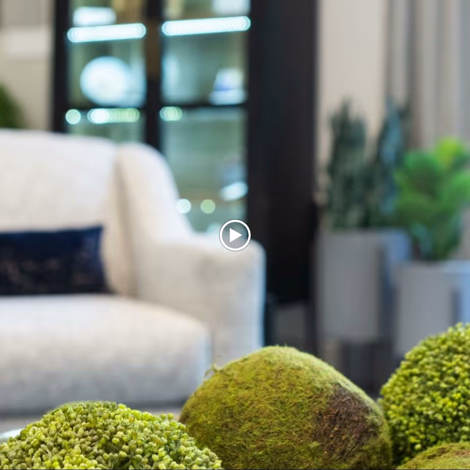 Close-up view of decorative green moss balls on a table with a blurred background showing a white upholstered armchair with a dark cushion and some potted plants in a modern living room setting.