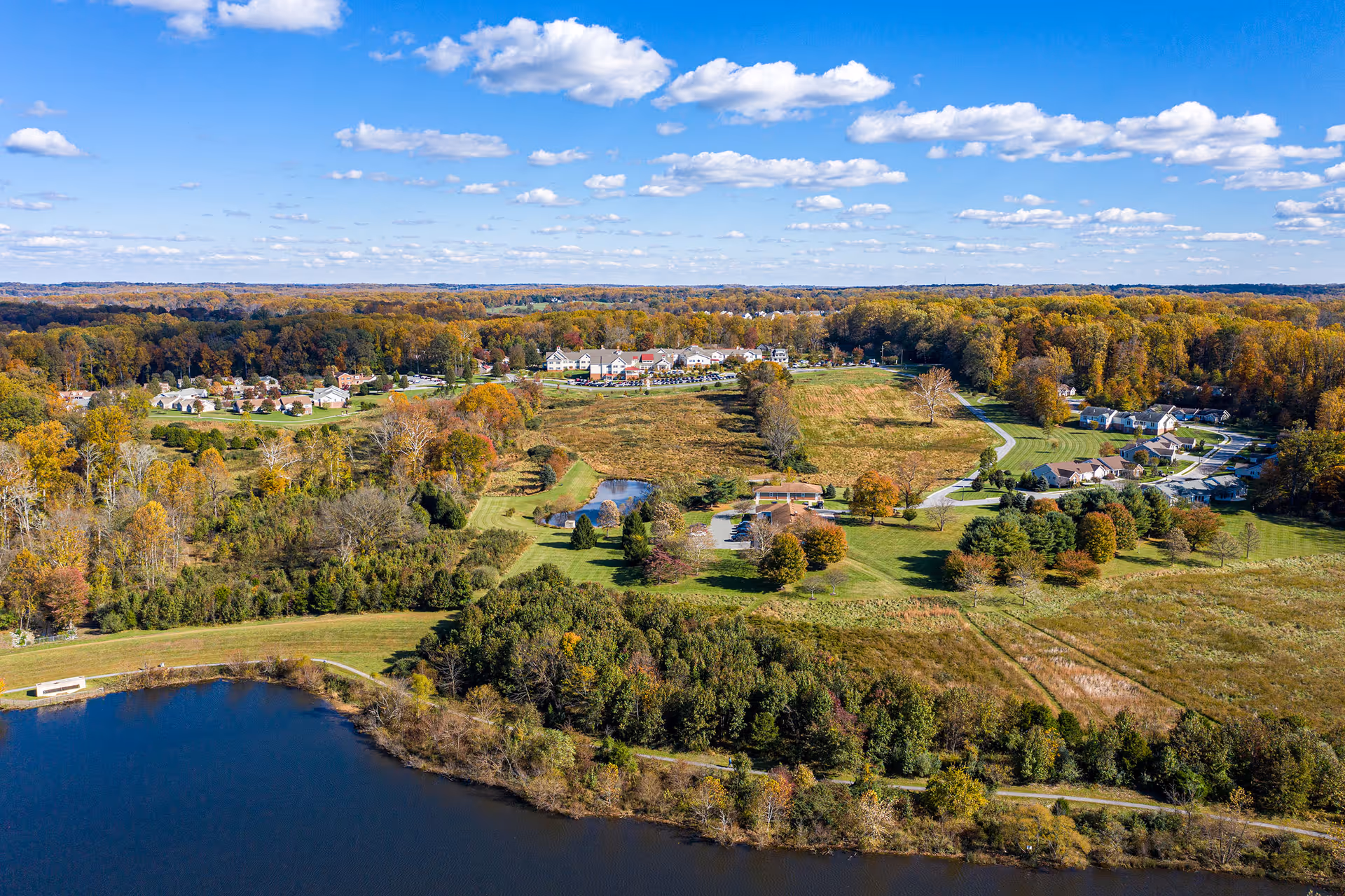 Aerial view of Brooke Grove Retirement Village showing a large body of water in the foreground, surrounded by trees and greenery. The retirement village buildings and houses are visible in the midground, with a mix of open grassy areas and wooded sections extending into the background under a partly cloudy blue sky.