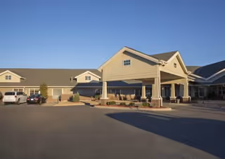 Front entrance of a single-story senior living facility with a covered porte-cochère, parked cars, landscaped shrubs, and a clear blue sky.